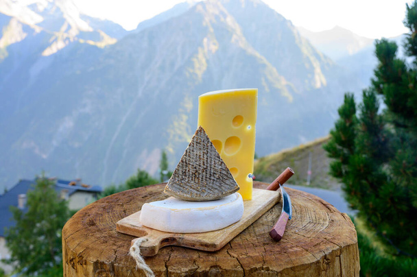 Käse-Kollektion, französischer Emmentaler, Tomme und Reblochon de Savoie Käse, der im Freien in der Region Savoyen serviert wird, mit alpinen Berggipfeln im Sommer im Hintergrund - Foto, Bild