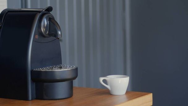 Zwarte compacte automatische Nespresso koffiezetapparaat in de buurt van een kleine witte keramische beker op een houten tafel. Hoge resolutie jpg afbeelding. - Foto, afbeelding