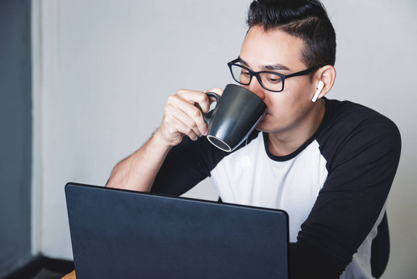 Fehér férfi teát iszik egy fekete bögréből, miközben otthon dolgozik, és a laptopja előtt ül. - Fotó, kép