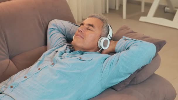 Odpočiňte si doma po práci během karantény. Starší muž s šedivými vlasy leží na pohovce a poslouchá hudbu pomocí bílých sluchátek nebo sluchátek. Volný čas. Koncept moderní technologie. - Záběry, video