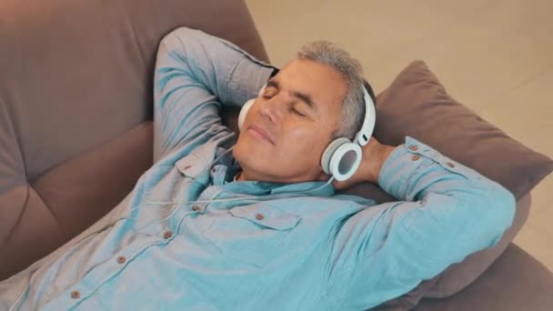 Pihenj otthon munka után a karantén alatt. Felnőtt férfi ősz hajjal fekszik a kanapén, és zenét hallgat fehér fülhallgatóval vagy fejhallgatóval. Szabadidő van. Modern technológiai koncepció. - Felvétel, videó