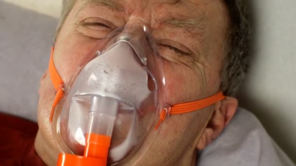 Zieke man van middelbare leeftijd die zwaar ademt door zuurstofmasker terwijl hij in bed ligt. Behandeling van astma, allergieën, bronchitis, longontsteking bij Covid-19 en respiratoire aandoeningen. Close-up. - Video
