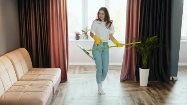 若い女性は家で掃除する。ブルネット女性モデルが踊っていると家を掃除しながらモップで歌っている. - 映像、動画