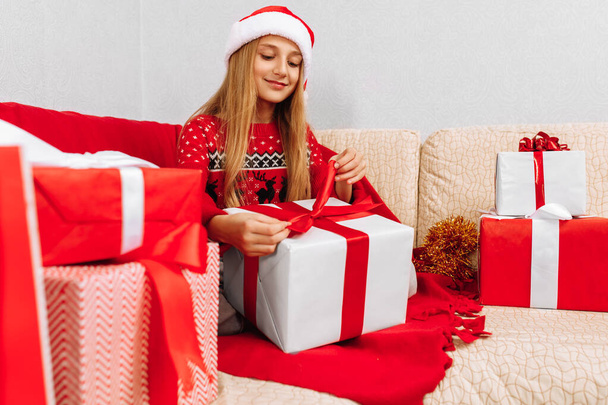 Happy smiling child wearing Santa Claus hat opening Christmas present while sitting on sofa at home, Merry Christmas and Happy Holidays - Fotoğraf, Görsel