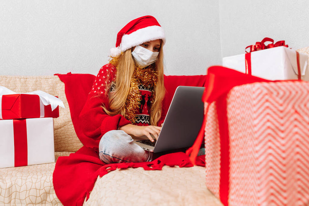 Little girl wearing medical mask, christmas sweater and santa claus hat while talking to online friend on laptop while sitting on sofa at home for christmas - Foto, Imagem