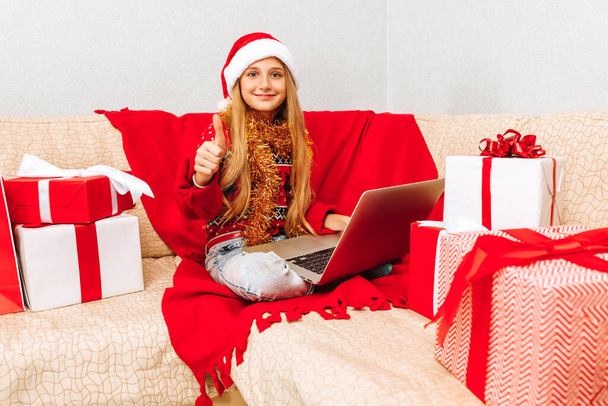 Little happy girl in Christmas Santa Claus hat sitting on sofa at home with laptop showing thumbs up - Foto, Imagem