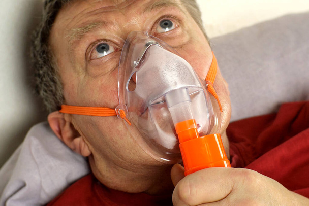 Άρρωστος ηλικιωμένος άντρας αναπνέει με μάσκα οξυγόνου, στριφογυρίζει τα μάτια του. Θεραπεία άσθµατος, αλλεργιών, βρογχίτιδας, πνευµονίας στο Covid-19 και αναπνευστικών νόσων. Επιλεκτική εστίαση. Κοντινό πλάνο. - Φωτογραφία, εικόνα