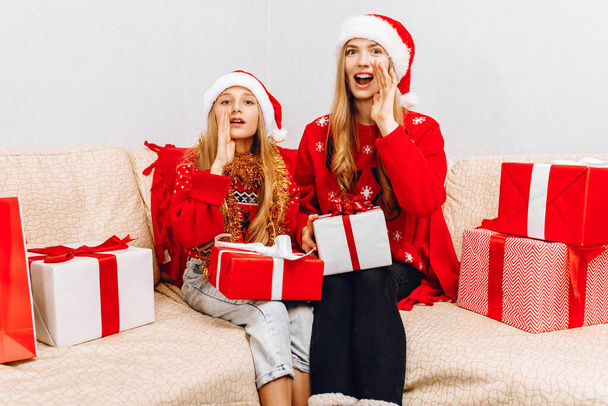 Happy family, beautiful mother and her little daughter in Santa Claus hats, are sitting at home on the couch with Christmas gifts, loving family with gifts, Merry Christmas and Happy New Year - Фото, изображение