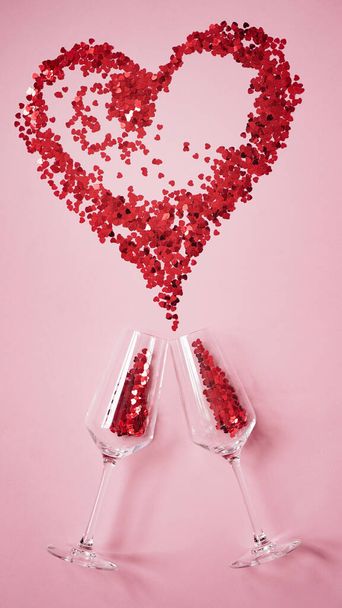 Two champagne glasses with red candies in shape of heart,big red heart made of candy as splash of champagne on pink background.Concept for greeting card,banner of valentine's day.Vertical photo - Photo, Image
