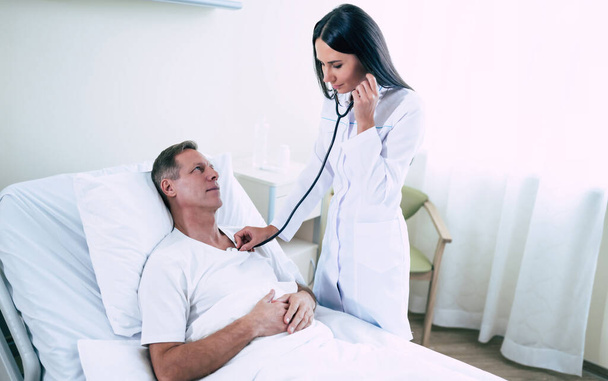 Young modern confident woman doctor in a white coat with a stethoscope is examining the patient's feelings while he is lying on the bed in the hospital ward. - Foto, Bild