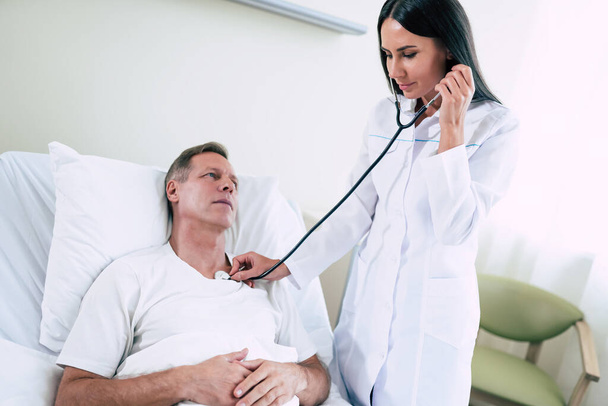 Young modern confident woman doctor in a white coat with a stethoscope is examining the patient's feelings while he is lying on the bed in the hospital ward. - 写真・画像