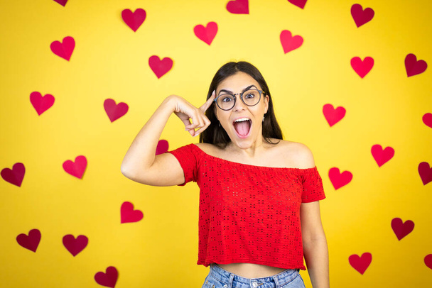 Young beautiful woman over yellow background with red hearts smiling and thinking with her fingers on her head that she has an idea. - Photo, Image