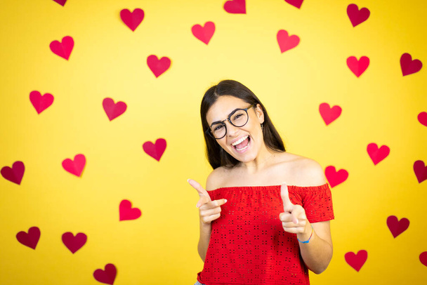 Молодая красивая женщина на желтом фоне с красными сердцами указывая на вас и камеру с пальцами, улыбаясь позитивно и весело - Фото, изображение