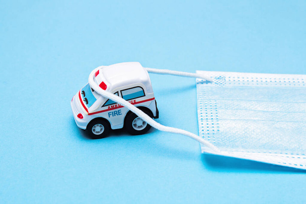 Toy ambulance car with white medical mask on the blue background. - Фото, изображение