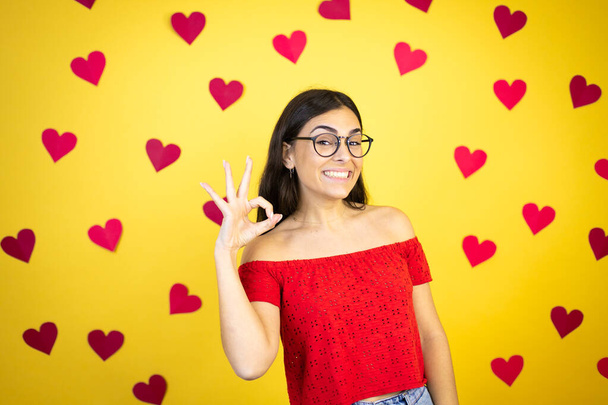 Jonge mooie vrouw over gele achtergrond met rode harten doen ok teken met vingers en glimlachen, uitstekend symbool - Foto, afbeelding