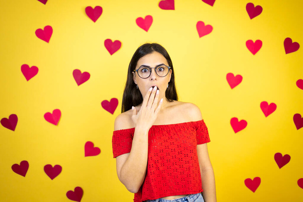 Young beautiful woman over yellow background with red hearts covering mouth with hand, shocked and afraid for mistake. surprised expression - 写真・画像