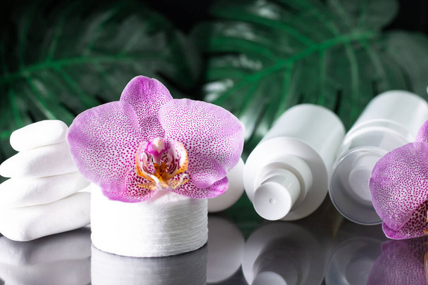 Hermosa flor de orquídea lila, botellas de cosméticos blancos, almohadillas de algodón con pilas de piedras blancas y hojas de monstera en la superficie negra. SPA o salón de belleza y concepto de procedimientos - Foto, Imagen