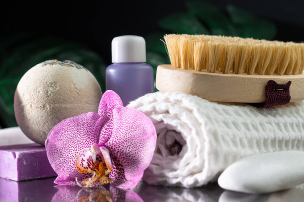 Beautiful lilac orchid flower, cosmetic bottle, massage brush, rolled towel and handmade soap with white stone and monstera leaves on black surface. SPA or beauty salon and procedures concept - Photo, Image