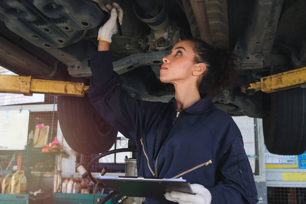 ガレージで働く女性の自動車整備士、自動車サービス技術者の女性は、自動車サービスセンターで顧客の車をチェックして修理し、身体とサスペンションシステムの下で車を検査し、車両修理サービスショップコンセプト. - 写真・画像