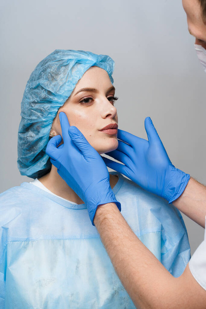 Consultation with plastic surgeon on white background. Cosmetic rejuvenating facial treatment. Doctor man with blue medical gloves touches girl face. Patient ask about lips and chin injections - Photo, Image