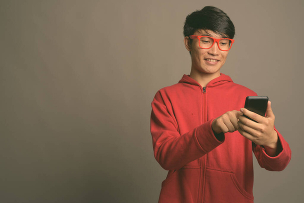 Молодой азиат в красной куртке в очках на сером фоне - Фото, изображение