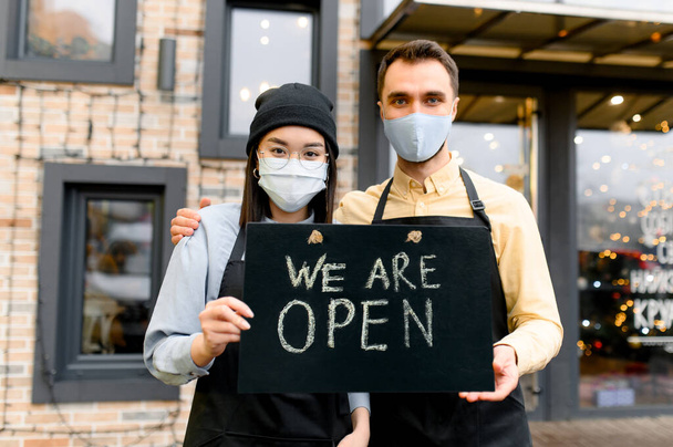 Welcome, we are OPEN. Two friendly waiters wearing protective masks. Multiracial waiters, caucasian guy and asian girl standing outside a restaurant, cafe or bar and showing signboard OPEN - Foto, Imagen
