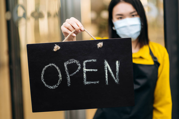 Signboard OPEN in the foreground. Defocused female waiter in a protective medical mask on her face, holds sign OPEN at the entrance to a restaurant or coffee shop. Small business concept - Foto, Imagem