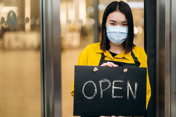 レストランやカフェの入り口に立っている幸せなウエイトレスの肖像画彼女の顔に医療マスクを着用。若いアジアの女性は彼女の小さなビジネスショップでオープンサインを示し、クライアントを招待 - 写真・画像