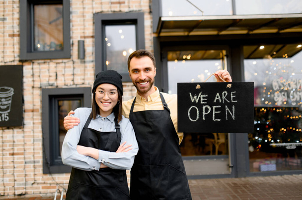 We are OPEN. Two multiracial friendly waiters wearing uniform stand outside a restaurant, cafe or bar, showing signboard OPEN, with pleasured smiling, welcome. Teamwork concept - Zdjęcie, obraz