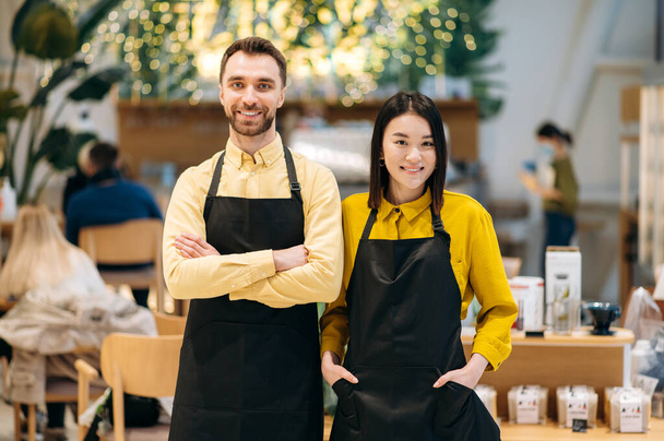 Praca zespołowa. Portret dwóch przyjaznych kelnerów. Atrakcyjny kaukaski facet i piękna azjatycka dziewczyna w czarnych fartuchach stoją wewnątrz restauracji, kawiarni lub baru i uśmiechnięty - Zdjęcie, obraz