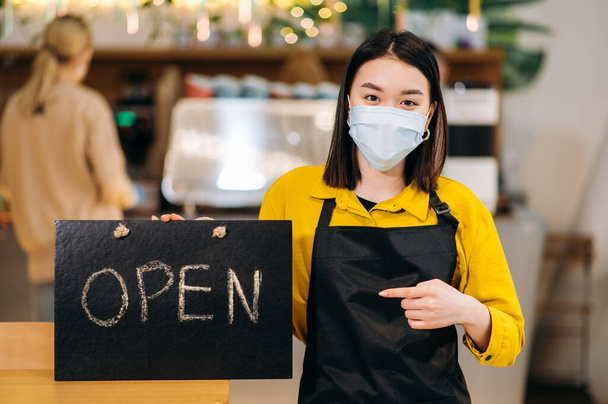 Signboard OPEN. Portrait of a young Asian waitress standing indoors of a restaurant or cafe wearing protective medical mask and black apron and holds signboard OPEN. Support small business concept - Photo, Image