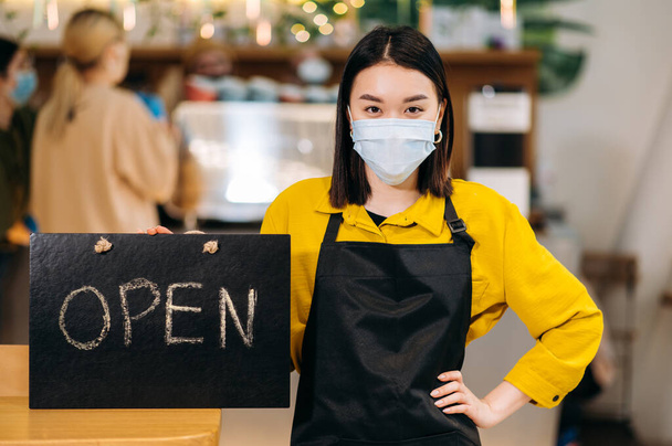 Welcome. Young Asian waitress stands indoors of a restaurant or cafe wearing protective medical mask and black apron and holds signboard OPEN. Support small business concept - Foto, afbeelding
