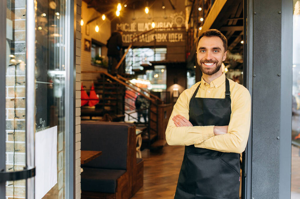 Portrait of an attractive bearded caucasian male waiter, barista or owner of a restaurant, cafe or bar stands at the entrance wearing a protective mask and a black work apron with crossed arms and - Photo, Image