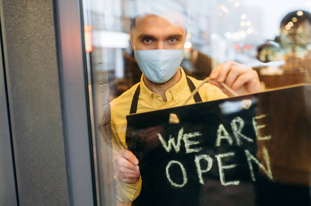 Welcome. Satisfied caucasian male waiter or barista showing signboard OPEN while standing at restaurant or cafe entrance wearing medical mask on his face and black apron and invites clients - Fotoğraf, Görsel