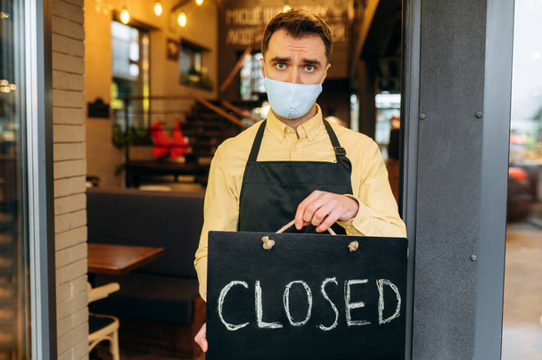 Smutný číšník, barista nebo malý podnikatel. Nešťastný muž v ochranné masce a černé pracovní zástěře stojí u vchodu do kavárny, restaurace nebo baru a je opatřen cedulí Uzavřeno - Fotografie, Obrázek