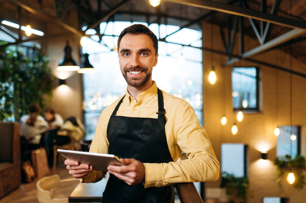 Ελκυστική ευτυχισμένη καυκάσιος σερβιτόρος ή barista φορώντας μαύρη ποδιά στέκεται μέσα σε ένα εστιατόριο, καφέ ή μπαρ, κατέχει ένα μενού και με φιλικό χαμόγελο κοιτάζει την κάμερα - Φωτογραφία, εικόνα