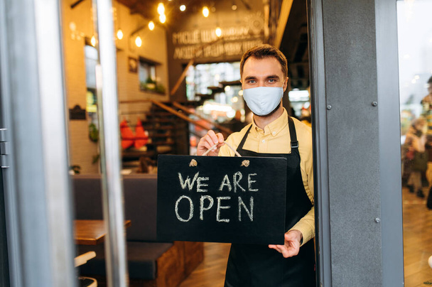 We are OPEN. Portrait of a satisfied caucasian waiter or barista standing at restaurant or cafe entrance wearing medical mask on his face and black apron. Employee man showing signboard OPEN in his - Foto, Imagen