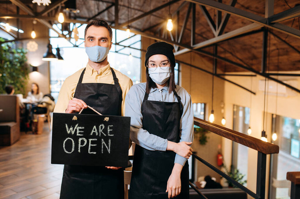 We are OPEN. Two friendly multiracial waiters wearing protective masks. Attractive caucasian guy and beautiful asian girl in black aprons stand inside a restaurant, cafe or bar and showing signboard - Photo, image