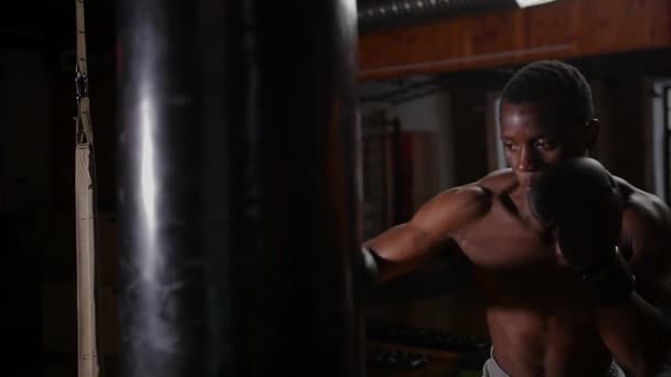Black shirtless man boxer training punching on the punching bag in the gym - Materiał filmowy, wideo