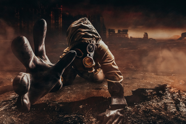 Foto de um perseguidor moribundo em jaqueta e luvas em máscara de gás danificada com filtro estendendo a mão para a câmera no fundo da cidade destruída deserto apocalíptico. - Foto, Imagem