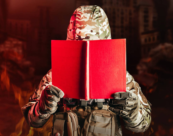 Foto di un soldato in uniforme e casco armatura tenuta aperto libro rosso su sfondo città in rovina. - Foto, immagini