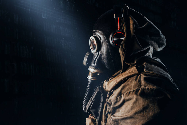 Photo of a stalker soldier in soviet rubber gas mask with  hose, wearing headphones and standing profile view on dark underground backdrop. - Photo, Image