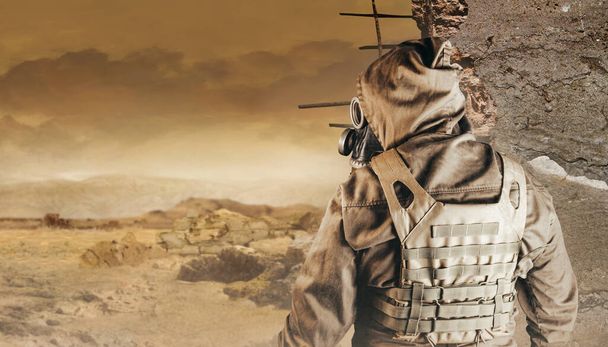 Photo of a stalker in jacket, armored vest standing back view in soviet gas mask with filter on destructed apocalyptic desert wasteland background. - Photo, Image