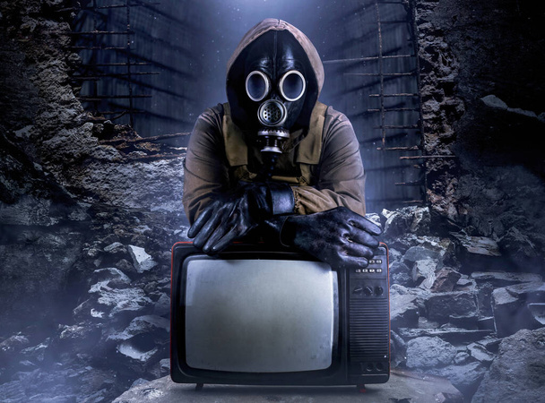 Photo of stalker soldier in jacket and armored vest and rubber gloves standing with old tv set on ruined dark background. - Photo, Image