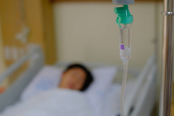 Imagen fuera de foco de una mujer que descansa en su cama de hospital después de una cirugía, durmiendo con una vía intravenosa goteando solución salina y medicamentos en la vena. - Foto, Imagen