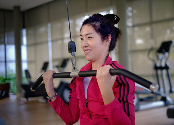 Een aantrekkelijke Aziatische vrouw is in het midden van haar training, gewichtheffen met behulp van een latissimus dorsi of een Lat pulldown machine om haar bovenlichaam te versterken in een sportschool. - Foto, afbeelding
