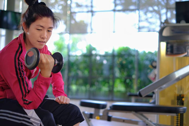 Привлекательная молодая азиатка, тренирующаяся в тренажерном зале, поднимающая вес гантелями, чувствующая усталость, но счастливая, улыбающаяся. - Фото, изображение