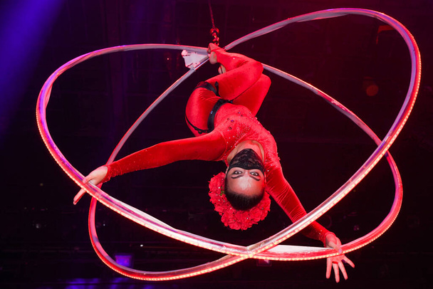 Flexible young woman make performance on aerial hoop, flexible back on aerial hoop, aerial circus show, blue and red light. Flexible woman gymnast upside down on hoop. Night club performance - Photo, Image