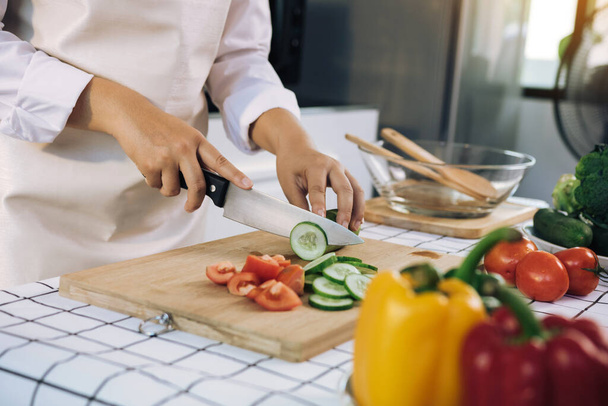 Frau schneidet mit Messer und Händen Gurke auf Holzbrett in Küche. - Foto, Bild
