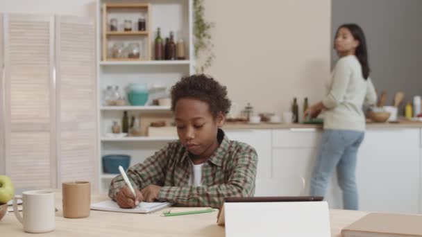 Peito de menino da escola Africano sentado à mesa em casa, escrevendo em caderno, olhando para computador tablet, mãe turva cozinhar e assistir filho na cozinha no fundo - Filmagem, Vídeo